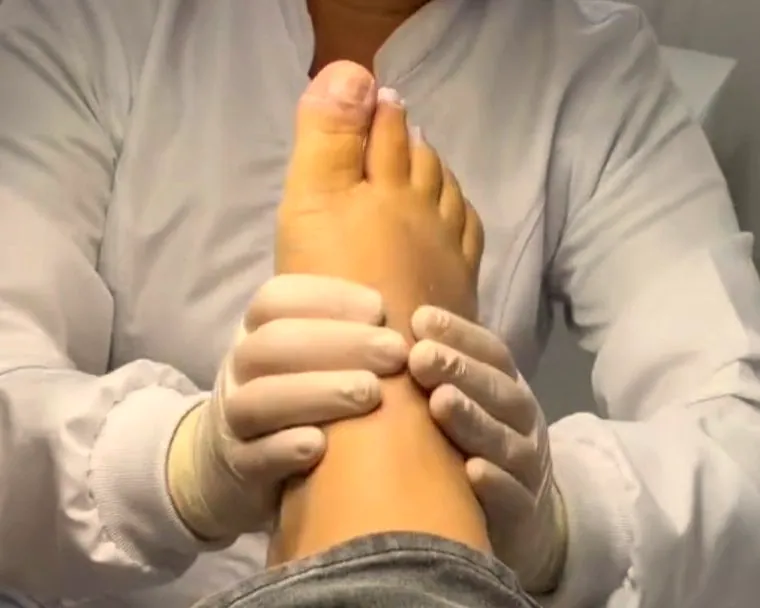 Mãos da podóloga, com luvas, segurando com cuidado o pé de uma cliente durante o atendimento